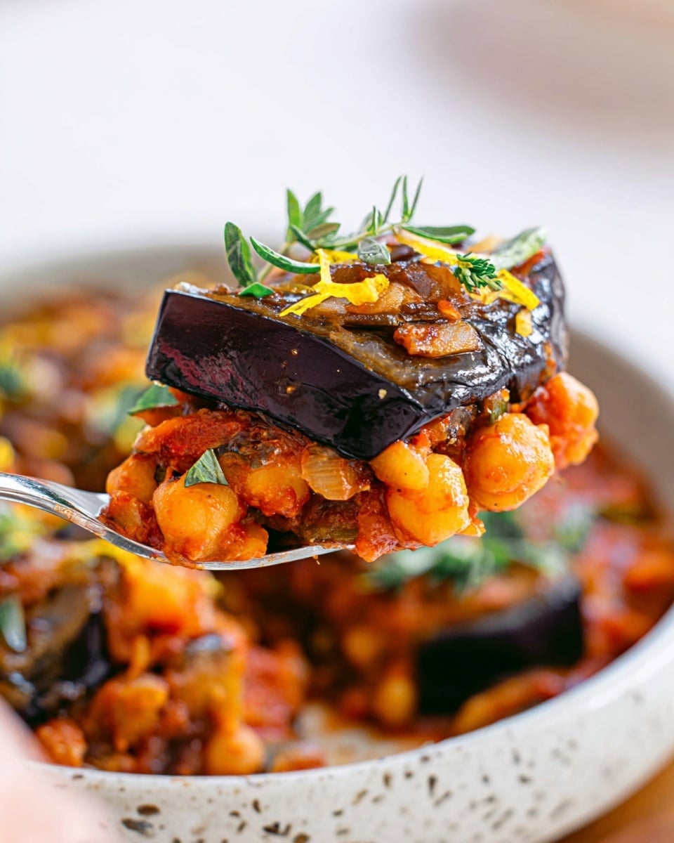 A close-up view of a cooked dish featuring two thick layers: the top layer is a shiny, soft, dark purple-black eggplant slice with a slightly oily surface, garnished with small green herb leaves and tiny yellow zest bits. Below it is a chunky, bright orange mixture consisting of chickpeas, diced onions, and tomato sauce pieces, adding a textured and moist base. The dish is served in a white marbled bowl, with more pieces of eggplant and the orange mixture visible beneath the main piece, creating a hearty and colorful combination. A silver spoon lifting a portion of the stack is held by a woman's hand. photo taken with an iphone --ar 4:5 --v 7