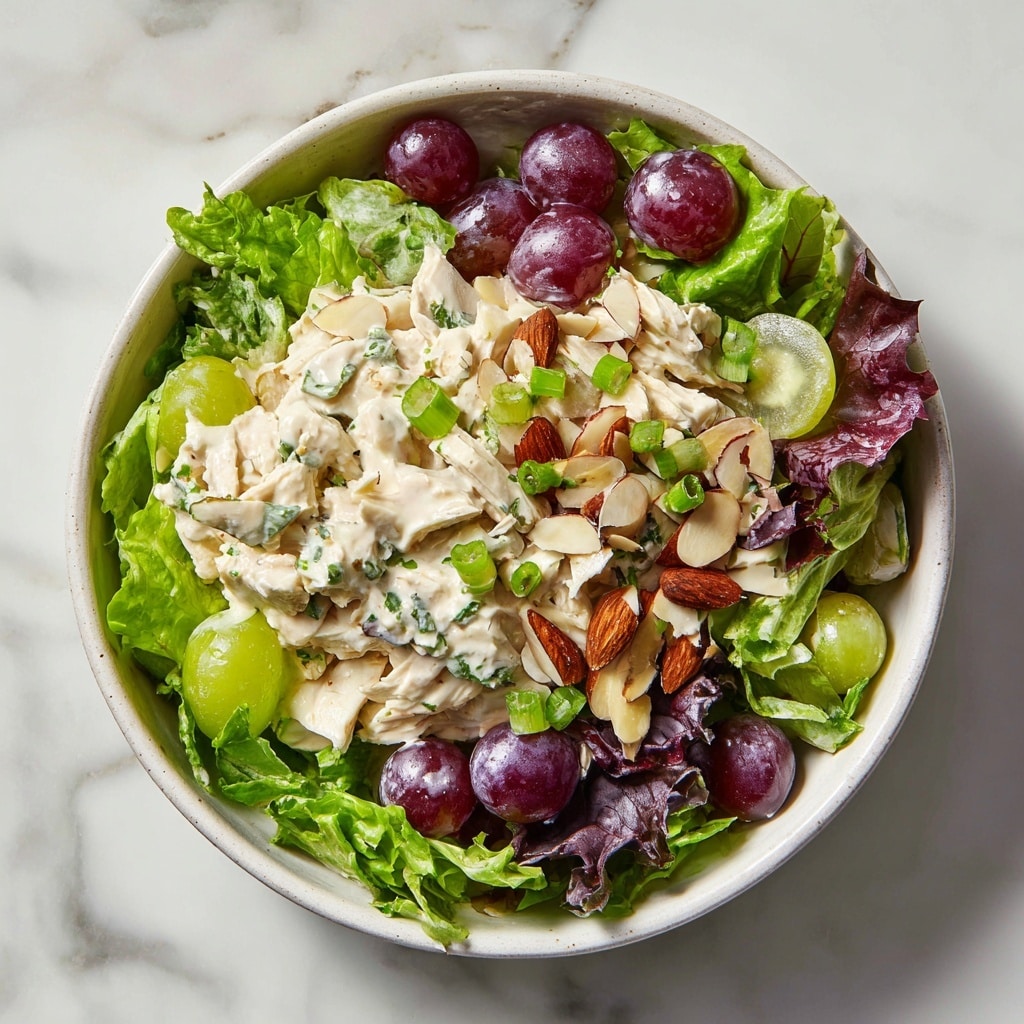 A bowl of chicken salad sits on a white marbled surface, featuring three main layers: the bottom layer is a mix of fresh green and purple leafy lettuce, the middle layer is creamy chicken salad with a light white sauce dotted with herbs, small chunks of celery, and sliced grapes both purple and green, and the top layer is scattered with sliced almonds and small pieces of green onion, adding texture and color contrast. The bowl is white with a simple, slightly textured rim. Photo taken with an iphone --ar 4:5 --v 7