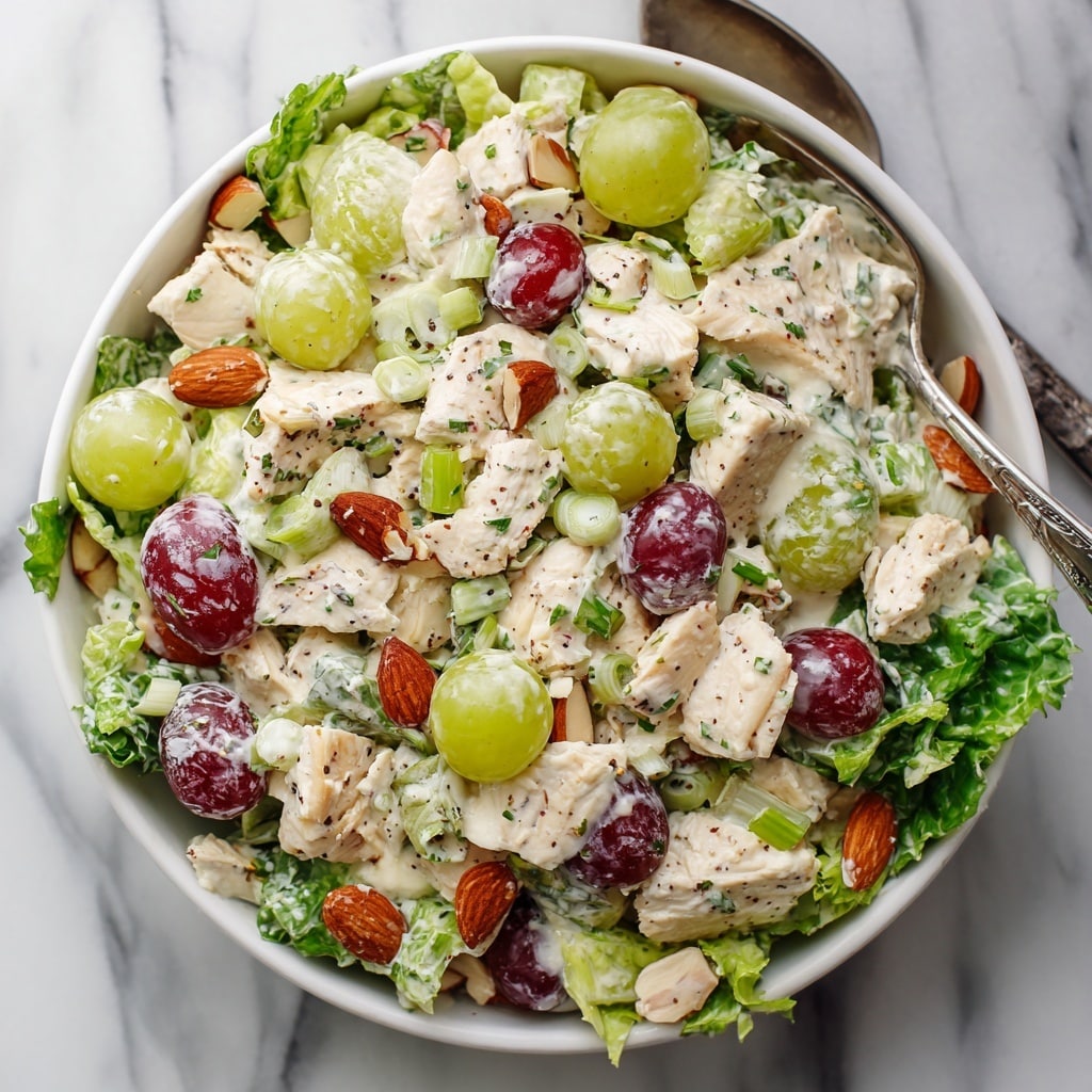 This image shows a white bowl filled with a creamy chicken salad on a white marbled surface. The salad has three main layers: the first layer is a base of fresh green leafy lettuce. The second layer sits on top, consisting of various ingredients including white chunks of cooked chicken, halved red and green grapes, pale green sliced celery, and scattered light brown almond slices. The third layer is a creamy dressing with a slightly grainy texture coating most ingredients, dotted with small green pieces of chopped green onions and herbs. A silver spoon is partially visible in the top right corner of the salad. The overall look is fresh with a mix of creamy, crunchy, and juicy textures. Photo taken with an iphone --ar 4:5 --v 7