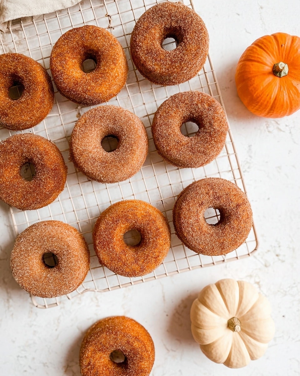 The image shows ten cinnamon sugar-coated donuts arranged on a white wire cooling rack with two donuts placed below it on a white marbled surface. Each donut is golden brown with a rough texture from the cinnamon sugar. On the right side of the cooling rack, there is a small bright orange pumpkin, and a pale cream pumpkin sits towards the lower right corner of the image. The overall setting gives a cozy fall feeling with warm colors and a clean background. photo taken with an iphone --ar 4:5 --v 7