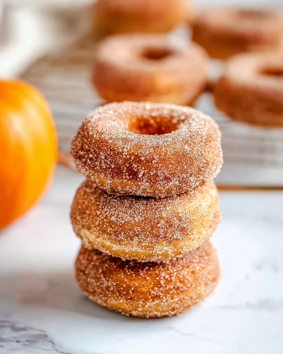 A close-up view of three round donuts stacked on top of each other, each covered in a light brown cinnamon sugar coating with a rough, grainy texture. The donuts have a golden-brown color beneath the sugar, showing a soft and slightly crisp surface. In the background, there are more donuts blurred out on a white cooling rack, and an orange pumpkin sits further back, all placed on a white marbled surface. The lighting is bright and natural, highlighting the sugar crystals on the donuts' surface. photo taken with an iphone --ar 4:5 --v 7