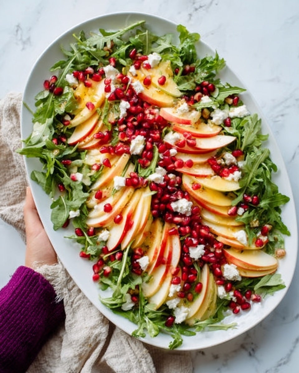The dish is a colorful salad served on a large white plate placed on a white marbled surface. The salad has several layers starting with a base of bright green arugula leaves spread evenly across the plate. On top, there are thin slices of red apple fanned out in small groups, adding a fresh red and yellow touch. Scattered over the salad are shiny, dark red pomegranate seeds that give a pop of deep color. Small bits of white cheese are sprinkled all over, adding a creamy texture. The whole dish looks vibrant and fresh, with a slight shine from a light dressing. Photo taken with an iphone --ar 4:5 --v 7