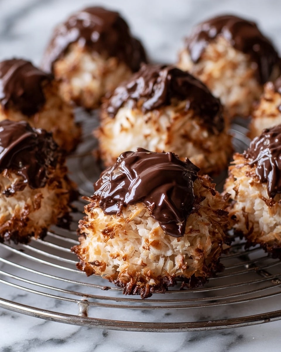 The image shows several small rounded mounds of toasted coconut covered in a dark glossy chocolate drizzle on top, arranged closely on a round metal cooling rack. Each mound has a coarse, rough texture from the shredded coconut, with the chocolate creating uneven shiny patches over the light and dark brown coconut. The background is a white marbled surface that softly blurs into a neutral light tone. photo taken with an iphone --ar 4:5 --v 7
