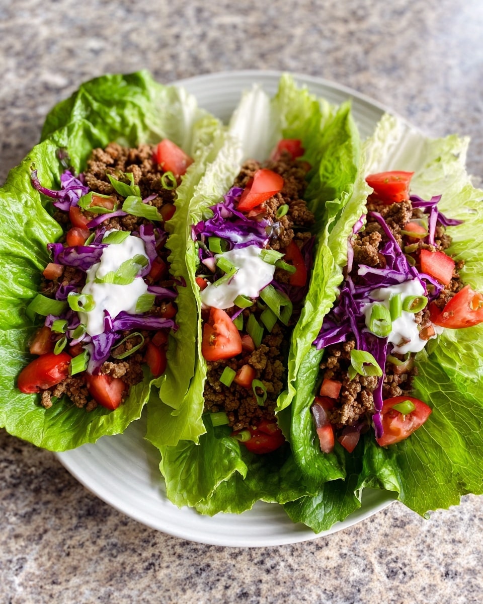 Three lettuce wraps are placed side by side on a white plate. Each wrap has a large green lettuce leaf as the base, topped with small pieces of purple cabbage, crumbled brown cooked meat, bright red chopped tomatoes, and a small dollop of white creamy sauce on top. Some green onion slices are scattered on the wraps as garnish. The plate sits on a white marbled textured surface. photo taken with an iphone --ar 4:5 --v 7
