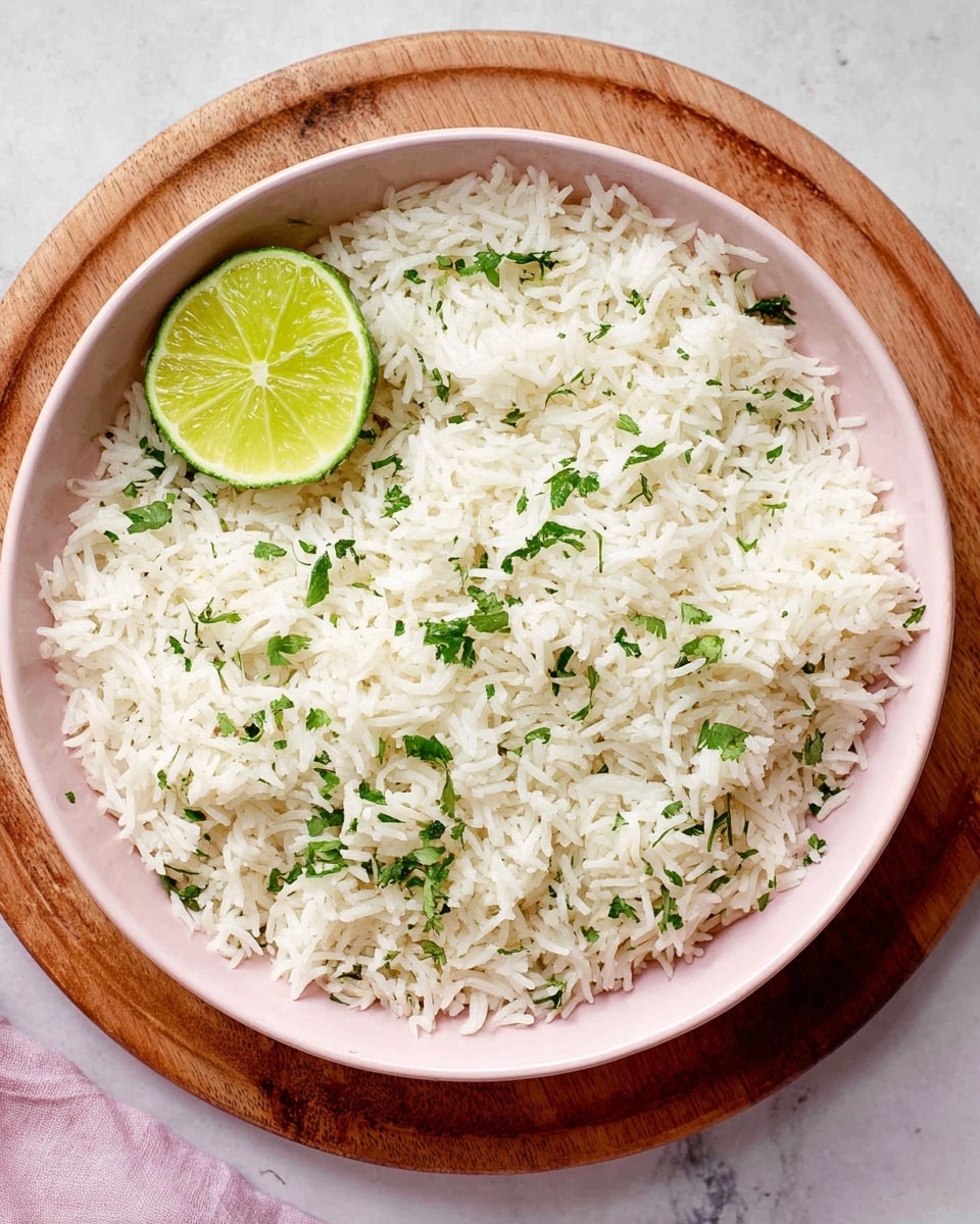 A pale pink bowl filled with a single layer of white rice mixed with small green cilantro pieces scattered evenly throughout. On the top left edge of the rice sits a quarter slice of lime with a bright yellow-green color. The bowl rests on a round wooden board, all placed on a white marbled textured surface. photo taken with an iphone --ar 4:5 --v 7