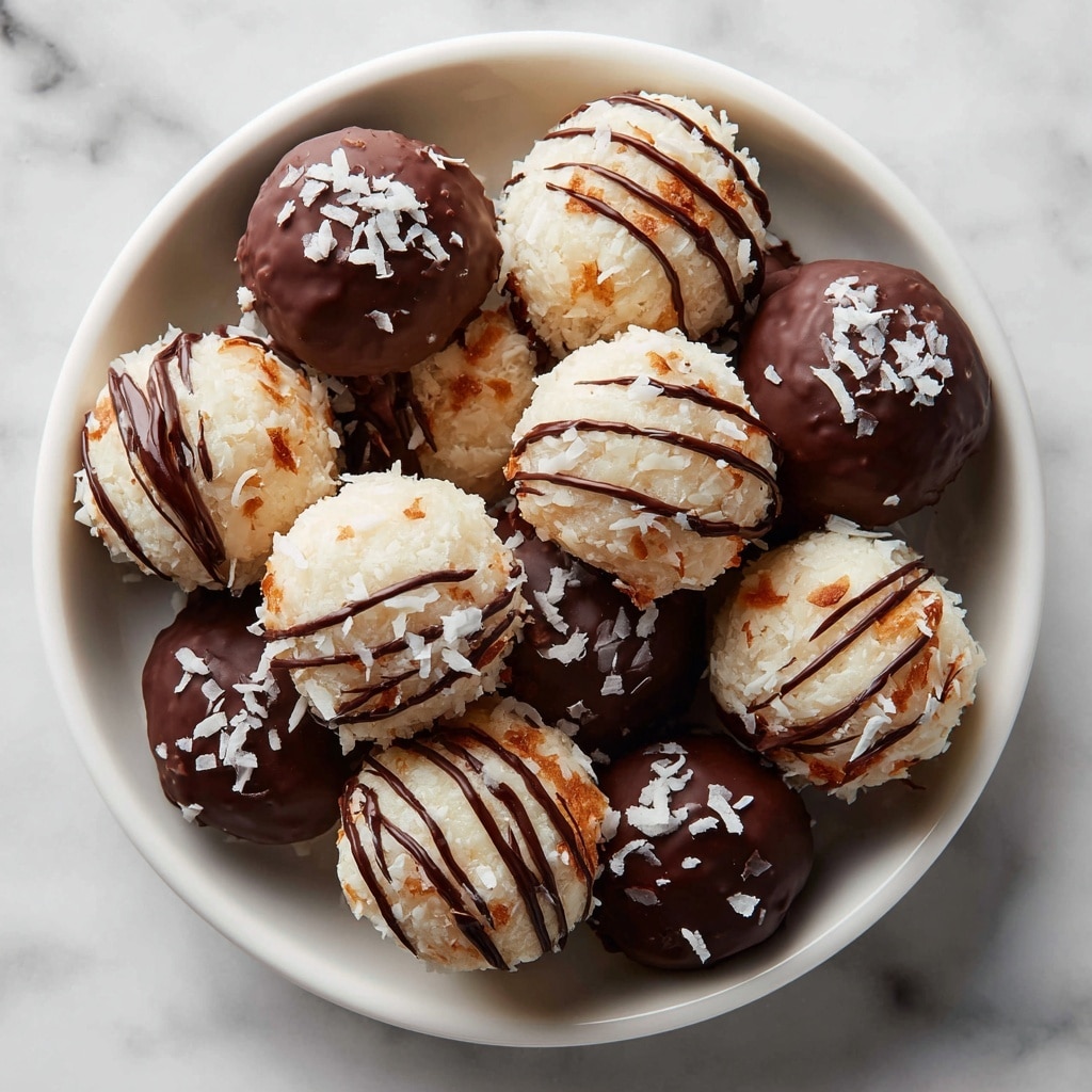 A white plate is filled with round coconut balls in two main styles: some are fully covered in smooth, dark chocolate with small white coconut flakes sprinkled on top, while others are white coconut balls drizzled with thin dark chocolate lines and also topped with coconut flakes. The coconut balls have a rough, textured surface from the coconut, contrasting with the smooth, shiny chocolate coating. The arrangement is close and piled, showing the mix of fully chocolate-covered and drizzled versions clearly. The background is a white marbled texture. Photo taken with an iphone --ar 4:5 --v 7