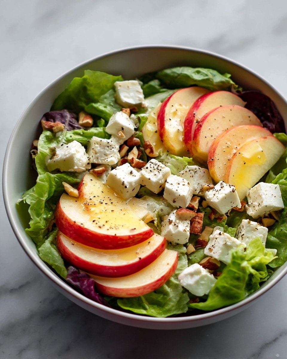 A white bowl holds a fresh salad starting with a base layer of green leafy lettuce, topped with several slices of red and yellow apple arranged in a fan shape. Scattered across the apples and lettuce are small white cubes of creamy cheese, likely feta, sprinkled with cracked black pepper. Small pieces of toasted nuts are also visible, adding texture and a crunchy contrast to the fresh ingredients. The salad has a light drizzle of oil, giving it a slight shine. The bowl is placed on a white marbled surface. photo taken with an iphone --ar 4:5 --v 7