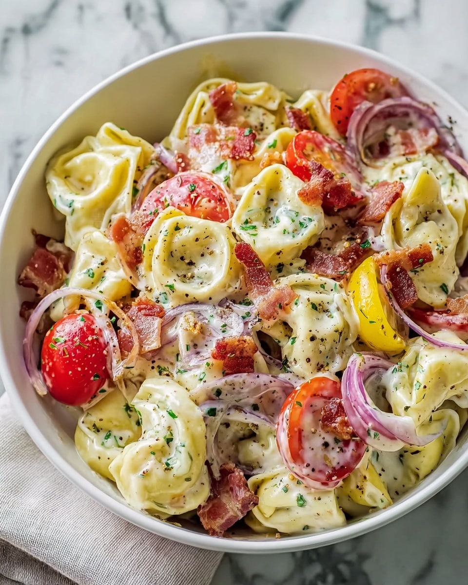 A white bowl filled with creamy tortellini pasta, each piece pale yellow with their folded shapes clearly visible. Mixed in are bright red cherry tomato halves, translucent purple rings of sliced red onion, and small pieces of crispy brown bacon scattered throughout. Thin slices of yellow pepper add a splash of color, and everything is coated with a creamy white sauce speckled with green herbs and black pepper. The bowl sits on a white marbled surface with a light-colored cloth partially showing beneath it. photo taken with an iphone --ar 4:5 --v 7