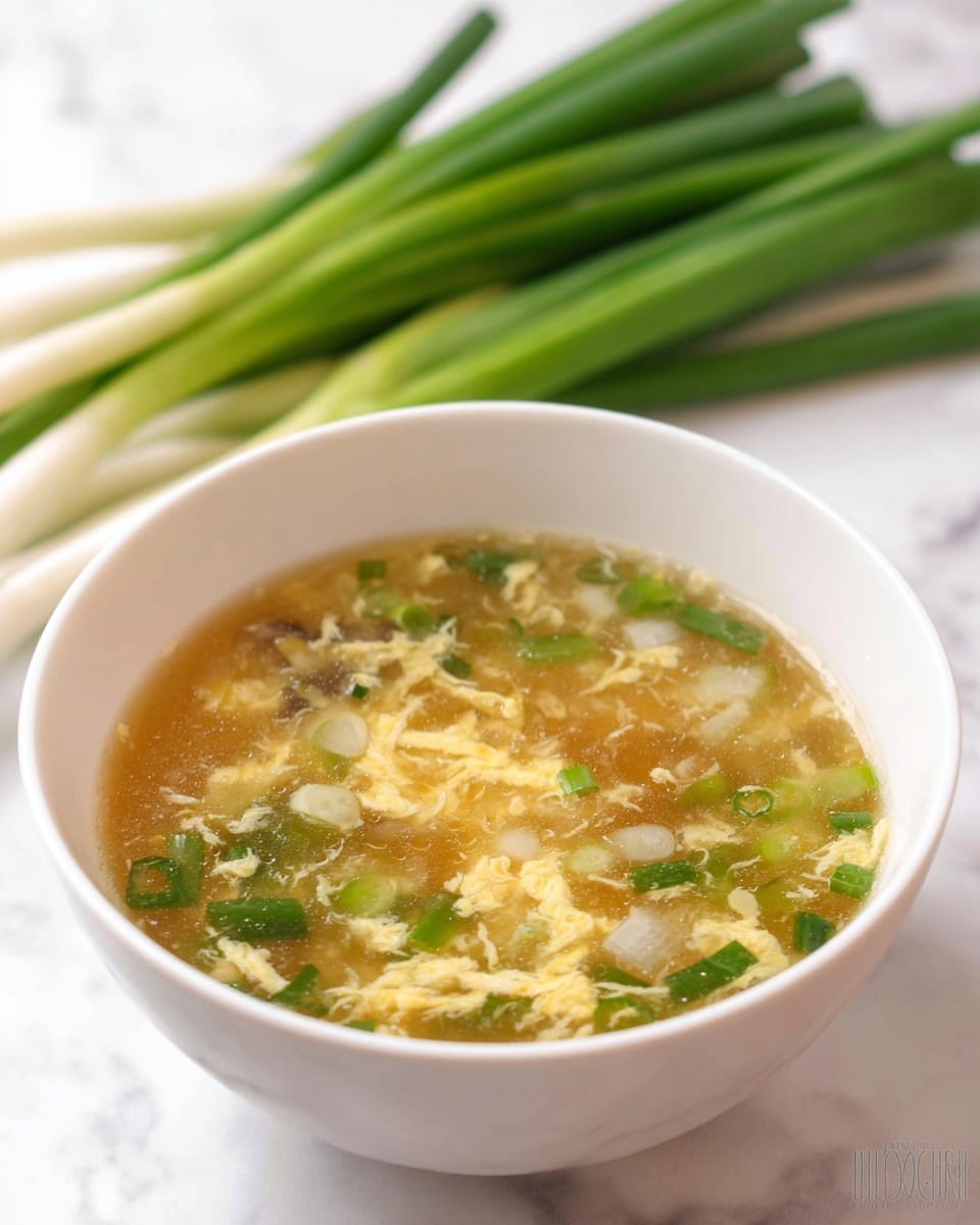 A white bowl filled with clear broth soup containing visible layers of light yellow, soft, shredded egg floating on top, mixed with green onion pieces and small chunks of translucent white vegetables, likely radish or mushroom slices. Behind the bowl, there are fresh whole green onions resting on a white marbled surface. The soup looks hot and comforting with steam lightly rising. Photo taken with an iphone --ar 4:5 --v 7
