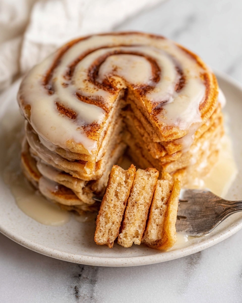 A stack of six thick, golden-brown pancakes sits on a white plate with smooth edges, each pancake showing a soft and fluffy texture with small air pockets. The top pancake is decorated with a spiral swirl of darker brown cinnamon, and a light glaze of creamy, white icing lightly drips over the edges. A bite-sized stack of three pancakes is held by a silver fork near the front right, revealing the multilayered inside with a warm, tan color and fluffy, slightly moist appearance. The plate rests on a surface with a white marbled texture. photo taken with an iphone --ar 4:5 --v 7