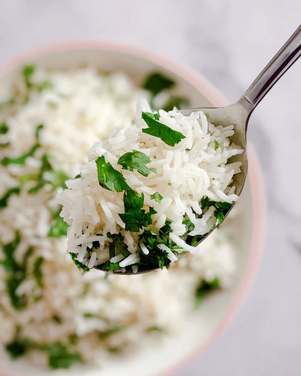 A close-up of a metal spoon holding a scoop of cooked white rice mixed with bright green cilantro leaves, showing the individual long grains and fresh herbs clearly. Below the spoon, there is a soft-focus view of a white plate filled with more rice and cilantro, resting on a white marbled texture. The image uses natural lighting to highlight the fresh, clean colors and slightly shiny texture of the rice grains and cilantro leaves. photo taken with an iphone --ar 4:5 --v 7
