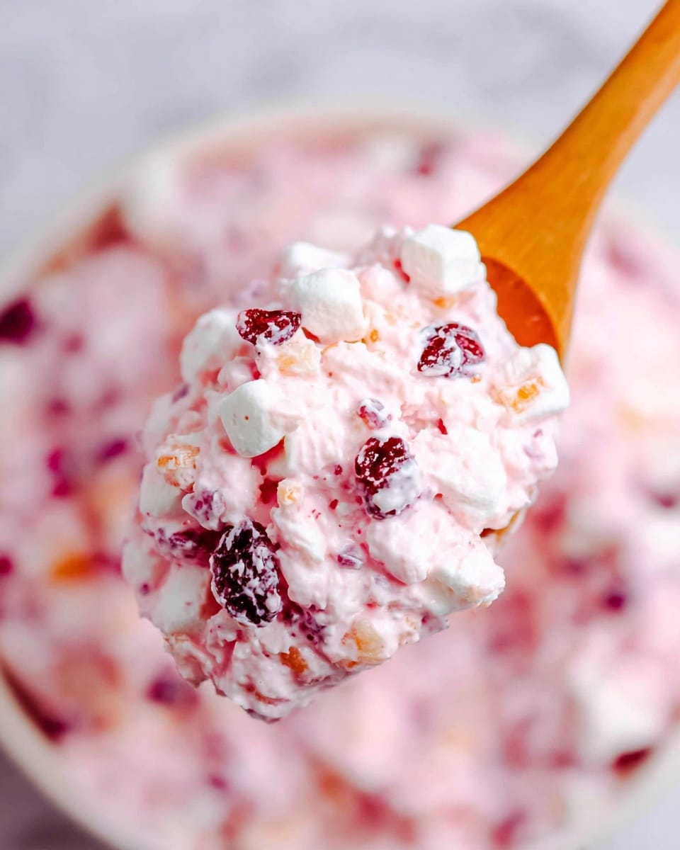 A close-up of a wooden spoon holding a scoop of a creamy pink salad with visible layers of small white marshmallows, dark red dried cranberries, and light orange fruit pieces, all mixed in a soft pink dressing; the background shows the same mixture in a white bowl with a white marbled surface beneath. photo taken with an iphone --ar 4:5 --v 7