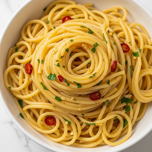 A white bowl filled with a large mound of spaghetti pasta tossed in olive oil with finely chopped green herbs and red chili flakes evenly spread throughout. The pasta strands are glossy and pale yellow, twisted loosely into layers that rise in the center, showing the texture of the herbs and chili mixed in. The simple, fresh ingredients highlight the smooth, slightly oily surface of the noodles against the clean white bowl, all set on a white marbled texture. photo taken with an iphone --ar 4:5 --v 7