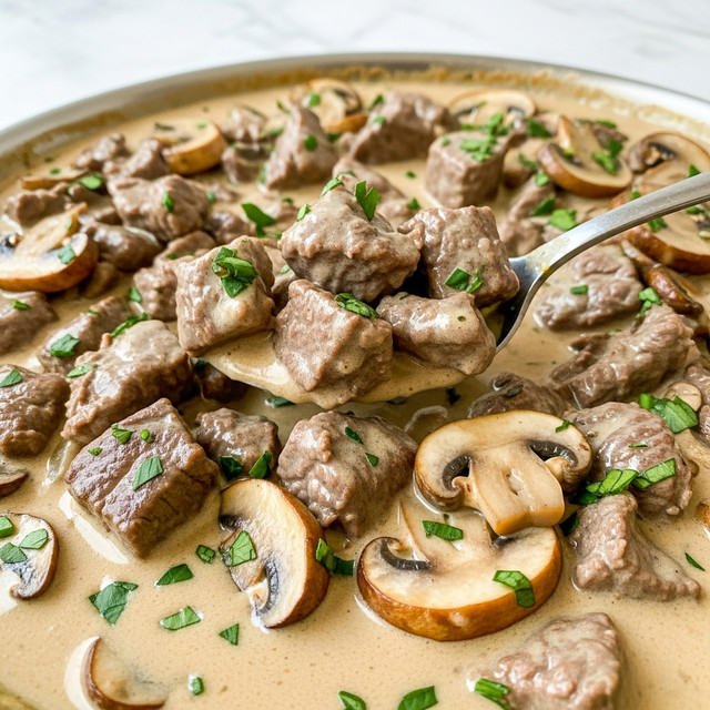 The image shows a creamy mushroom and beef stew in a pan, with chunks of tender brown beef and sliced light brown mushrooms covered in a thick, light beige sauce. The stew is sprinkled with small, bright green chopped herbs, adding a touch of color. A silver spoon lifts a portion from the pan, showing a close-up of the smooth sauce, beef pieces, and mushroom slices in it. The pan sits on a white marbled surface. photo taken with an iphone --ar 4:5 --v 7