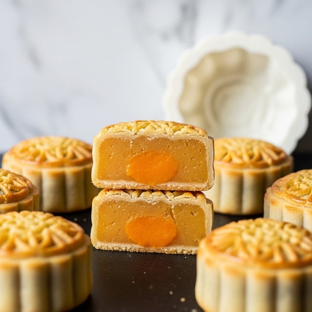 The image shows a close-up of a round mooncake cut in half, stacked one piece on top of the other. The outer crust is light golden yellow, with an ornate pattern on top, and has a soft, crumbly texture. Inside there are two thick layers of bright orange filling with a smooth, slightly glossy surface and a firm but soft consistency. In the background, more whole mooncakes and a white mooncake mold are visible on a white marbled surface, creating a cozy bakery feel. photo taken with an iphone --ar 4:5 --v 7