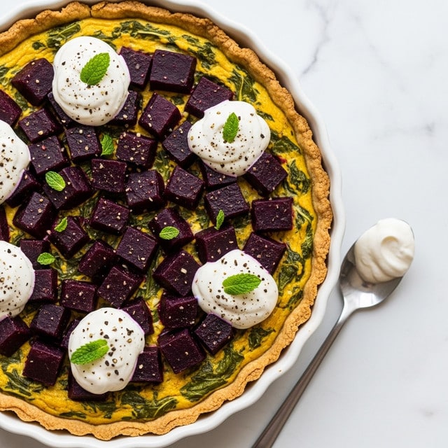 A round tart with a golden-brown crust holds a thick, creamy yellow filling mixed with green leafy pieces. Large, deep purple beet cubes are evenly layered on top, some partially sunk into the filling. Dollops of white creamy topping are spread around the tart's surface, each sprinkled lightly with black pepper and decorated with small green mint leaves. The tart sits in a white fluted dish placed on a white marbled texture surface, with a spoon beside it that has some of the white topping on it. photo taken with an iphone --ar 4:5 --v 7