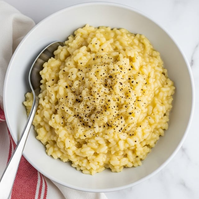 A white bowl filled with creamy risotto made of short, plump rice grains that look soft and moist. The risotto has a pale yellow color with a smooth, slightly glossy texture. The top is sprinkled with small black pepper flakes for a touch of contrast and texture. A silver spoon is placed inside the bowl, resting on the left edge. The bowl sits on a white marbled surface with part of a red and white cloth visible on the lower left side. photo taken with an iphone --ar 4:5 --v 7