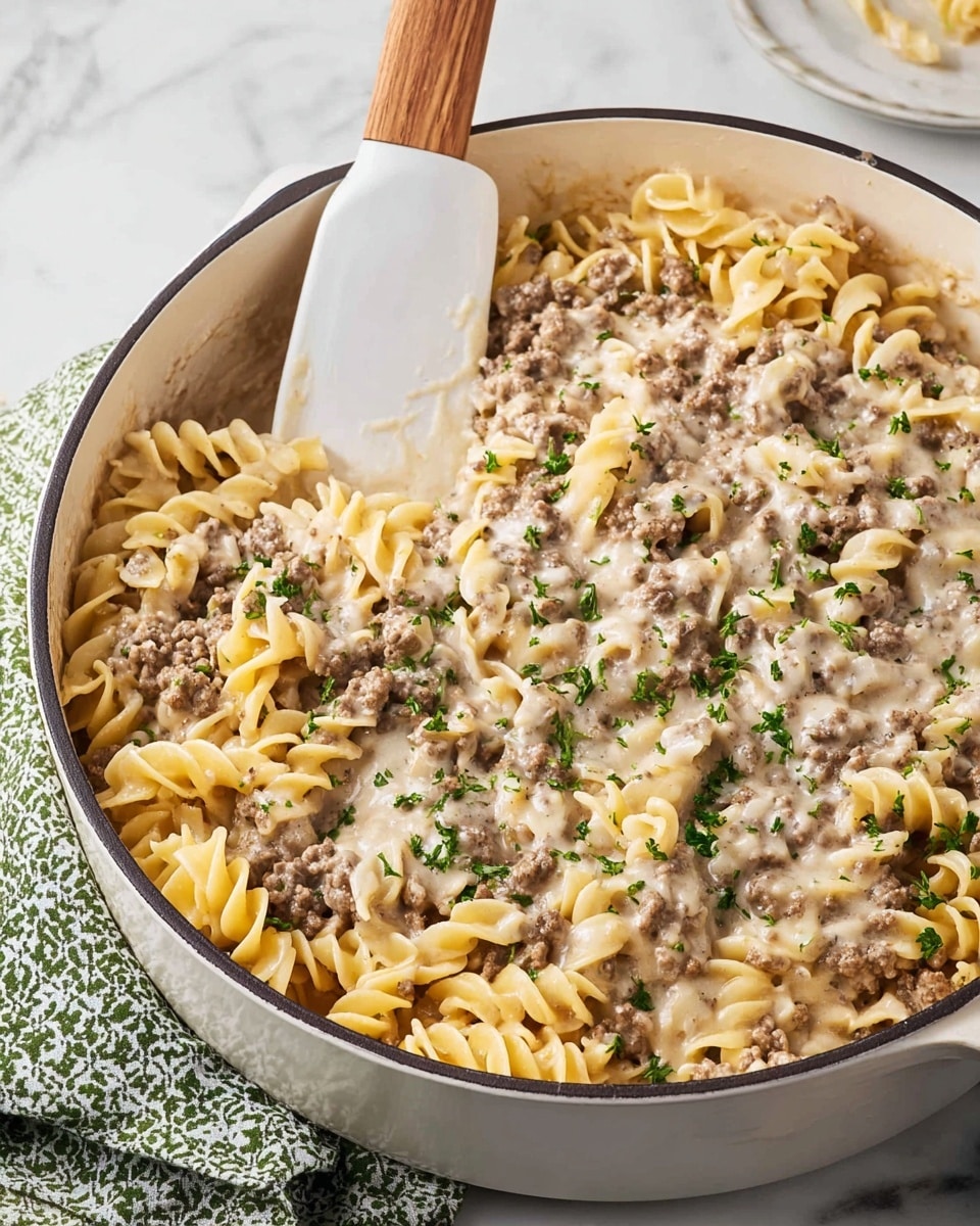 A white skillet filled with a creamy ground beef and egg noodle pasta dish, where tender, curly beige noodles are mixed evenly with small browned beef pieces in a light grayish creamy sauce, sprinkled with small bright green herb bits on top, a white spatula with a wooden handle is resting inside the skillet lifting some noodles, the skillet sits on a white marbled surface with a blurry salt shaker near the top right and a white and green patterned cloth at the bottom left, photo taken with an iphone --ar 4:5 --v 7