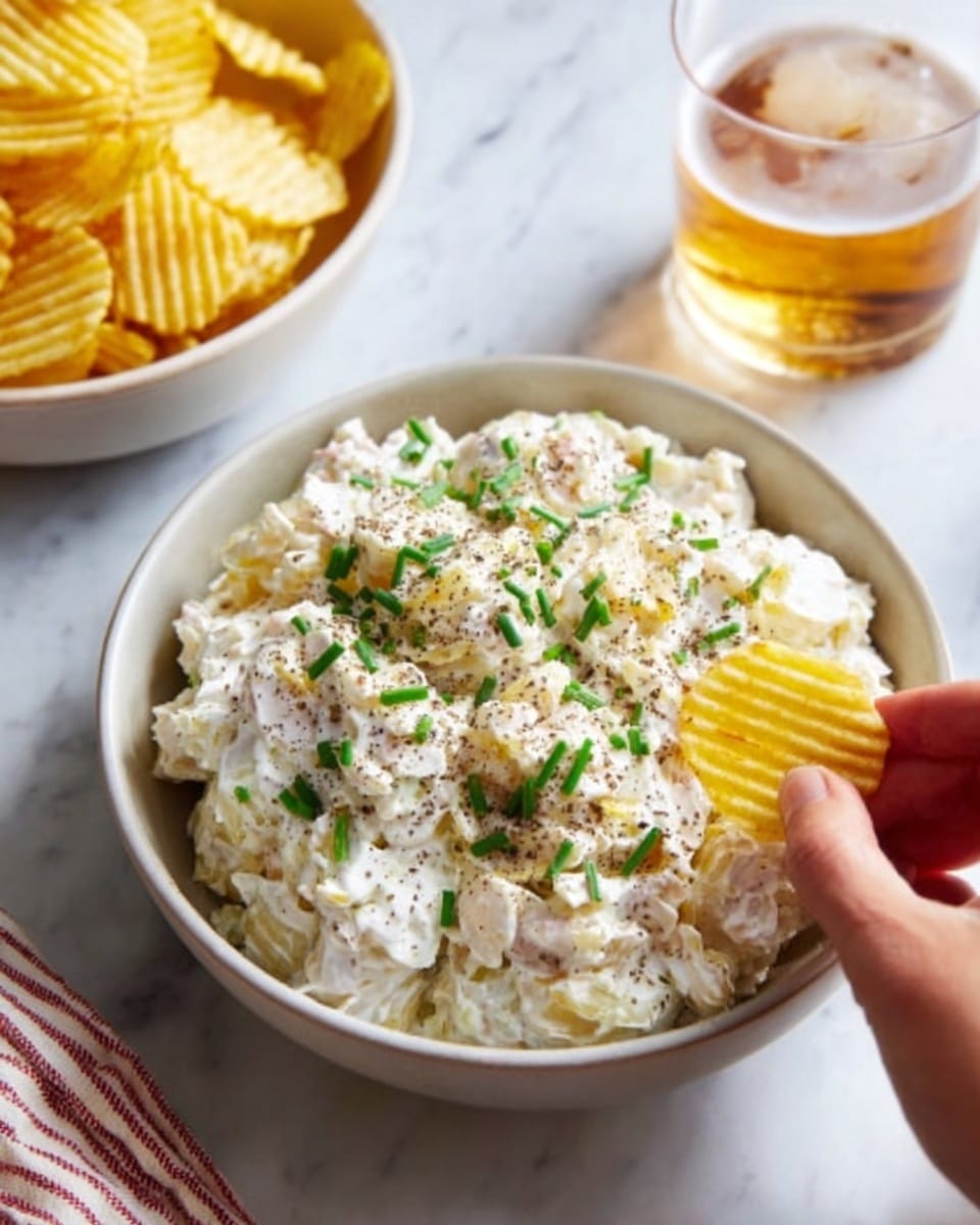 A white bowl filled with creamy potato salad topped with chopped chives and black pepper sits on a white marbled surface. The potato salad has several chunky potato pieces mixed with a thick, white mayo dressing, showing a smooth and slightly lumpy texture. In front of the bowl, a woman's hand holds a yellow ridged potato chip about to dip into the salad. Nearby is a white bowl filled with more yellow ridged potato chips and a glass of light amber-colored drink. The setting has a casual, inviting feel. photo taken with an iphone --ar 4:5 --v 7