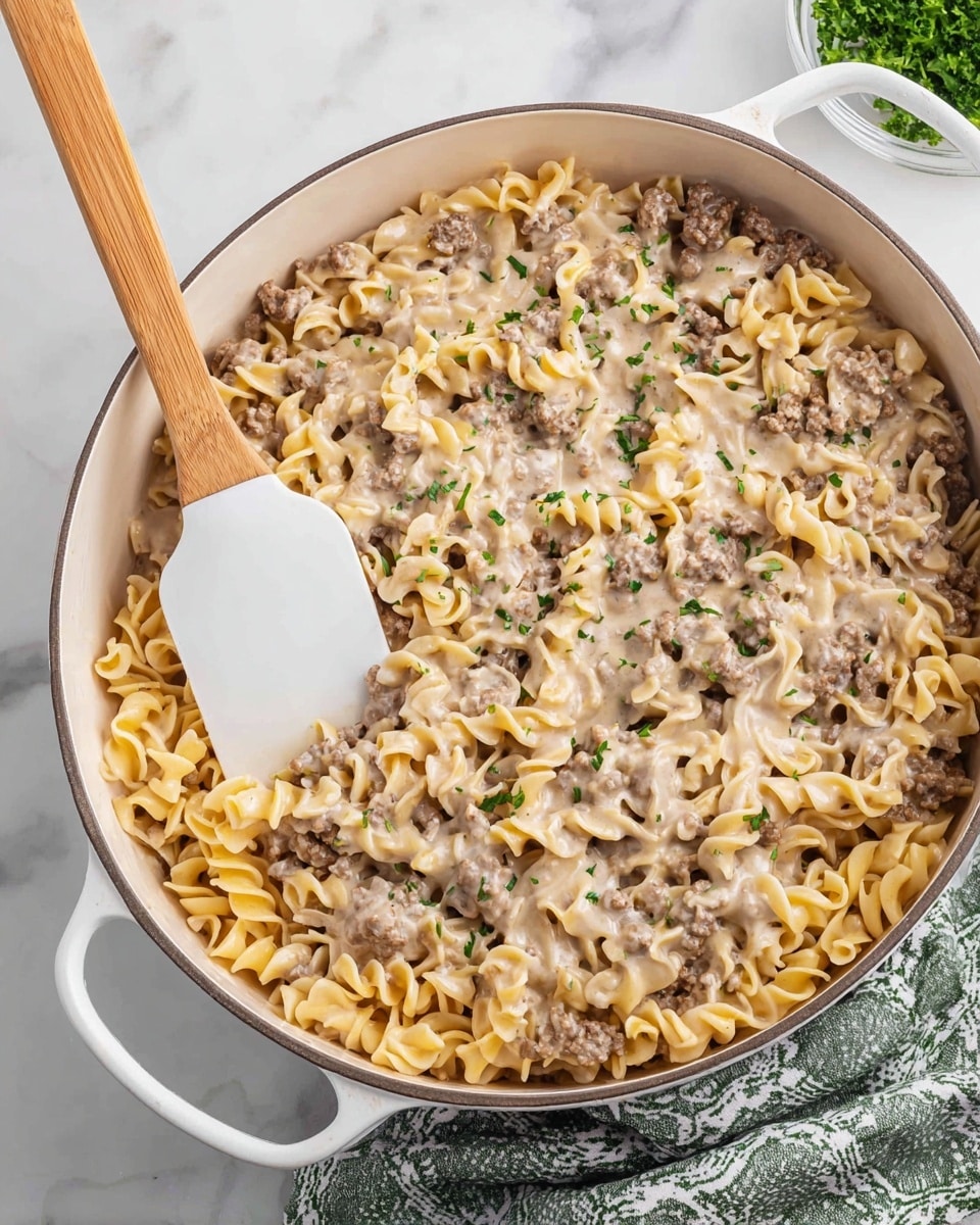 A white pan filled with creamy beef noodle casserole, showing three main layers: the bottom layer of soft, twisted pale yellow noodles, the middle layer of browned ground beef mixed through, and the top layer of smooth, light beige creamy sauce that coats everything evenly, with small green parsley pieces sprinkled across for color contrast; a white spatula with a wooden handle rests inside the pan, partially lifting some of the casserole, all placed on a white marbled surface with a green and white patterned cloth nearby. photo taken with an iphone --ar 4:5 --v 7
