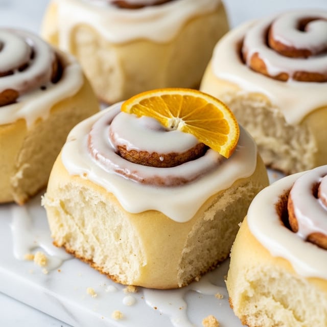 A close-up view of several soft cinnamon rolls each with one thick layer of creamy white icing that smoothly covers the top and part of the sides. The rolls have a light golden-brown baked dough with visible swirls of cinnamon inside. Some rolls are topped with a small fresh bright orange slice placed on top, adding a pop of color. They are all placed on a white marbled surface. The texture of the icing looks smooth and slightly glossy. Photo taken with an iphone --ar 4:5 --v 7