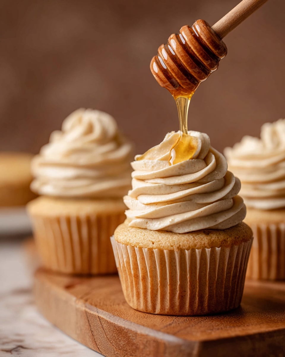 A close-up of three cupcakes on a wooden stand, each with one layer of light brown cake and a thick swirl of creamy off-white frosting on top, with visible small specks in the frosting. Above the closest cupcake, a wooden honey dipper drips golden honey onto the frosting, showing a shiny, smooth texture. The background is a soft blur with a white marbled texture, including a blurred honey bottle in the distance. photo taken with an iphone --ar 4:5 --v 7