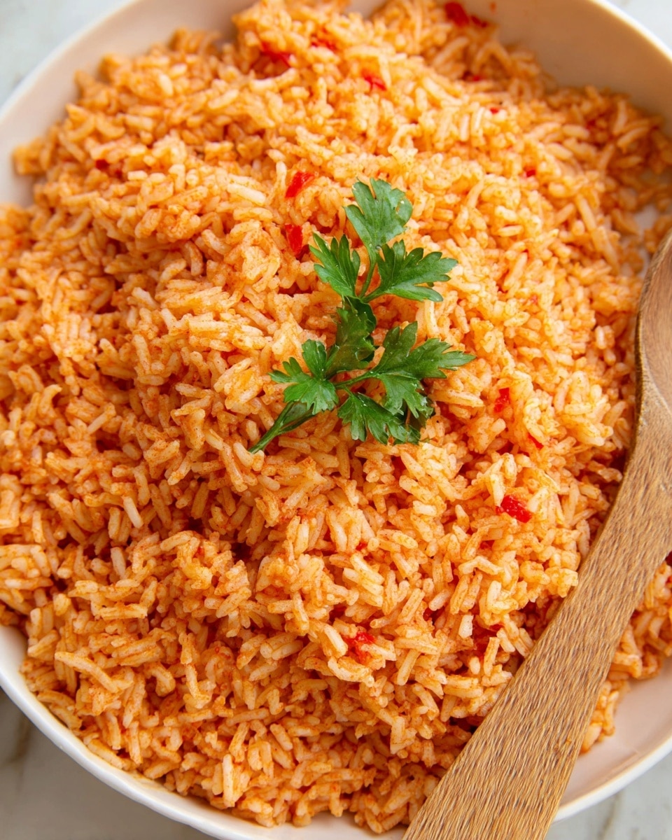 A close-up view of a single layer of cooked rice mixed with red sauce spreading evenly, giving orange and light red shades throughout; the texture looks soft and slightly fluffy, with some grains clumped together. On top at the center, a small green parsley sprig sits fresh, adding a touch of contrast. A wooden spoon is placed on the right edge of the white bowl, partially in the rice. The bowl is set against a white marbled texture as the background. photo taken with an iphone --ar 4:5 --v 7