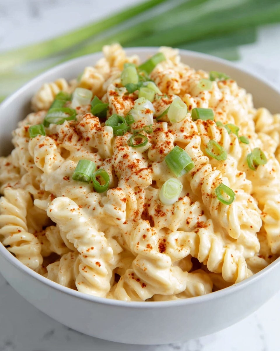 A close-up of a white bowl filled with creamy macaroni pasta mixed in a smooth, pale yellow cheese sauce, topped with scattered bright green chopped scallions, and a light dusting of reddish paprika powder evenly spread on top. The pasta pieces are layered naturally in the bowl, showing their curved and curly textures, with the sauce coating each piece richly. The background and surface show a soft white marbled texture. photo taken with an iphone --ar 4:5 --v 7
