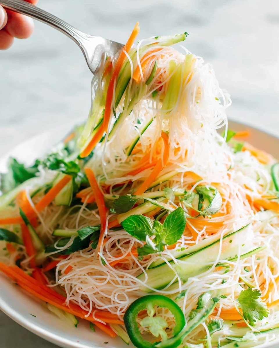 This image shows a white plate filled with a light and fresh noodle salad made of thin, transparent noodles as the base layer. On top, there are thin, long strips of bright orange carrots and pale green cucumber slices creating a crunchy texture. Green slices of jalapeño with visible seeds are scattered on the dish, adding a spicy touch. Fresh, leafy herbs like cilantro and basil, rich green in color, are spread unevenly over the noodles, giving a natural and fresh look. A spoon is lifting a portion of the noodles and vegetables, showing their delicate and mixed textures clearly, resting on a clean white marbled surface. Photo taken with an iphone --ar 4:5 --v 7