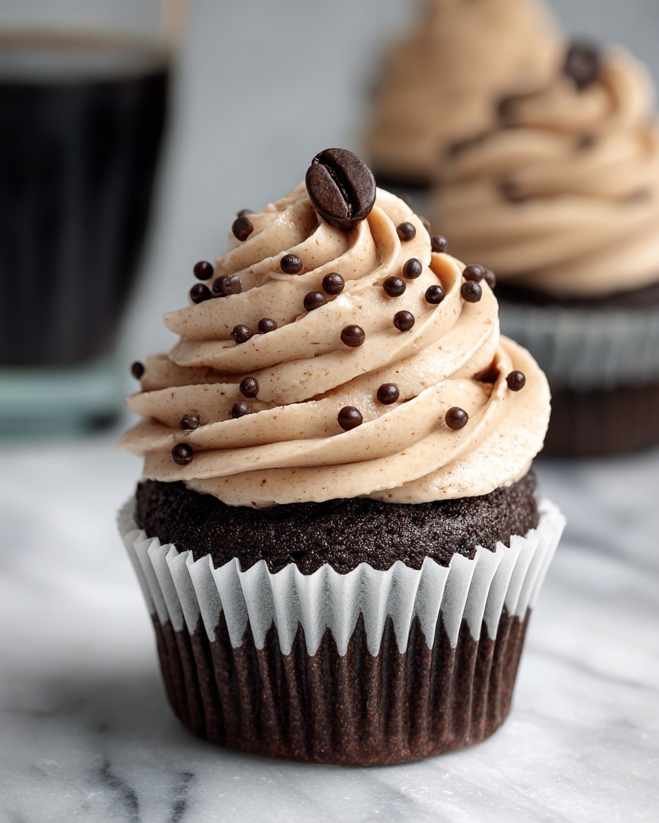A close-up of a single chocolate cupcake with a dark brown base in a white paper liner. On top, there is a thick swirl of light brown creamy frosting with a smooth texture, decorated with small round dark chocolate sprinkles evenly spread. At the very top of the frosting, a single dark brown coffee bean is placed. The cupcake sits on a white marbled surface with a blurred background showing part of another cupcake and a dark glass. A woman's hand reaches slightly from the side towards the cupcake. Photo taken with an iphone --ar 4:5 --v 7