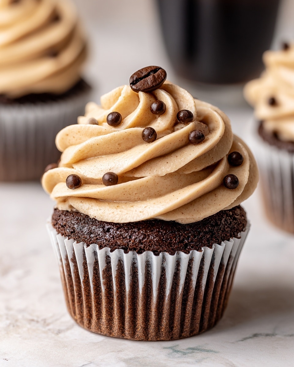 A close-up of a chocolate cupcake with three visible layers: a rich dark brown chocolate cake base, a white paper cupcake liner around the cake, and a thick swirl of light brown coffee-flavored frosting on top, decorated with small dark chocolate chips and a single glossy coffee bean at the peak. The cupcake sits on a white marbled surface with blurred background elements that include a glass and another cupcake. A woman's hand is partially visible, gently touching the cupcake. Photo taken with an iphone --ar 4:5 --v 7