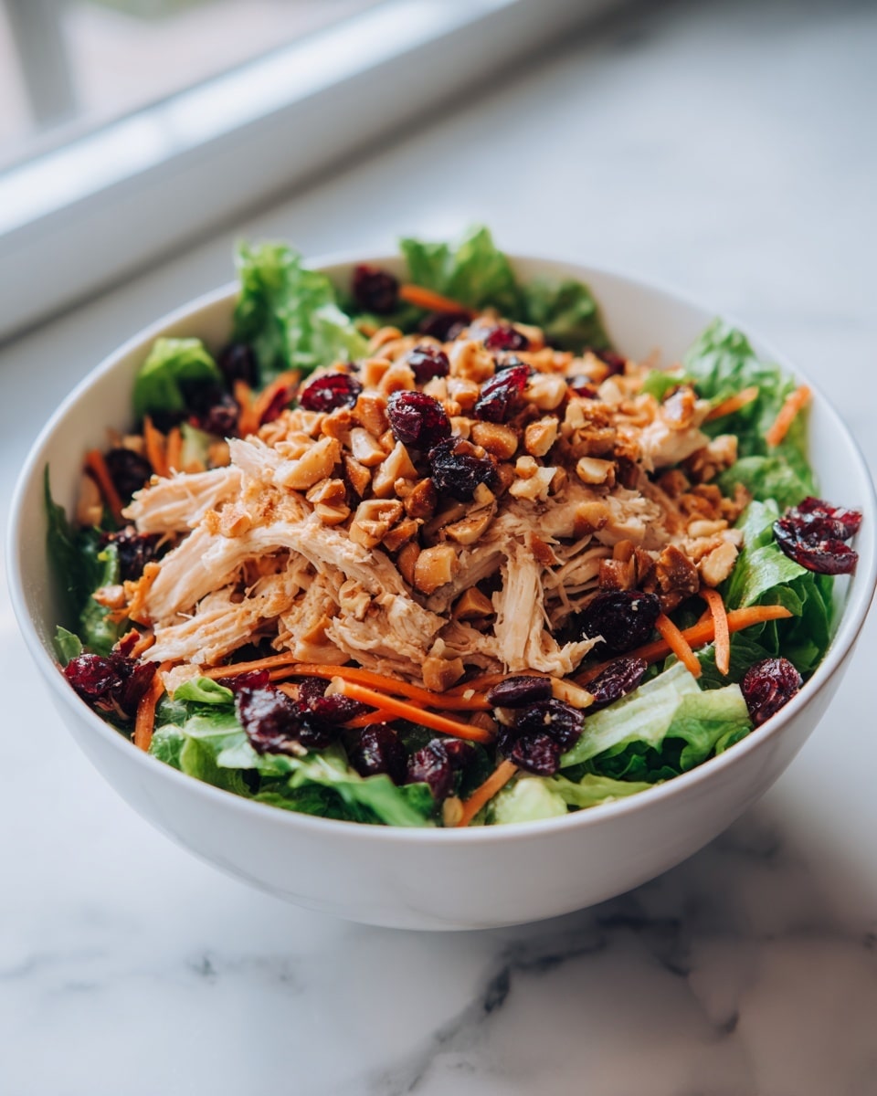 A white bowl filled with a fresh salad, starting with a base layer of bright green leafy lettuce. On top of the lettuce, there is a thick layer of shredded, light tan chicken. Scattered throughout the chicken are small, dark reddish-purple cranberries and thin, light orange carrot strips. The salad is finished with a generous topping of chopped, golden-brown nuts that add a crunchy texture. The bowl sits on a white marbled surface with soft daylight coming from the side. photo taken with an iphone --ar 4:5 --v 7