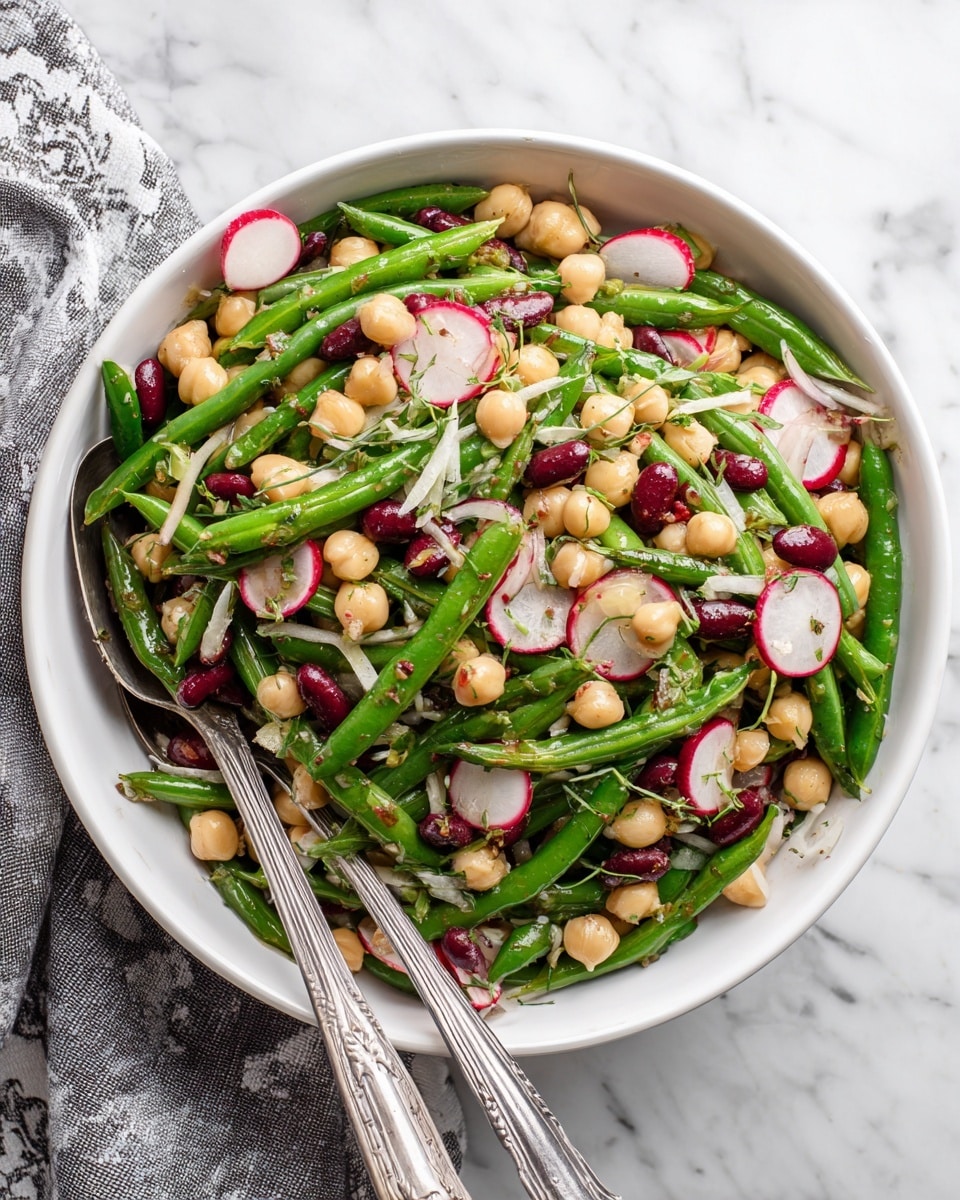 A close-up view of a large white bowl filled with a colorful bean salad, consisting of three main layers: bright green cut green beans, small round beige chickpeas, and dark red kidney beans mixed throughout. Thin white strips of onion and slices of radish with red edges and white centers are scattered on top, adding texture and contrast. The salad appears lightly coated with a dressing, adding a slight shine, and is garnished with small sprigs of fresh herbs. Two silver serving spoons rest inside the bowl. The bowl is placed on a white marbled surface with a grey patterned cloth partially visible on the side. Photo taken with an iphone --ar 4:5 --v 7