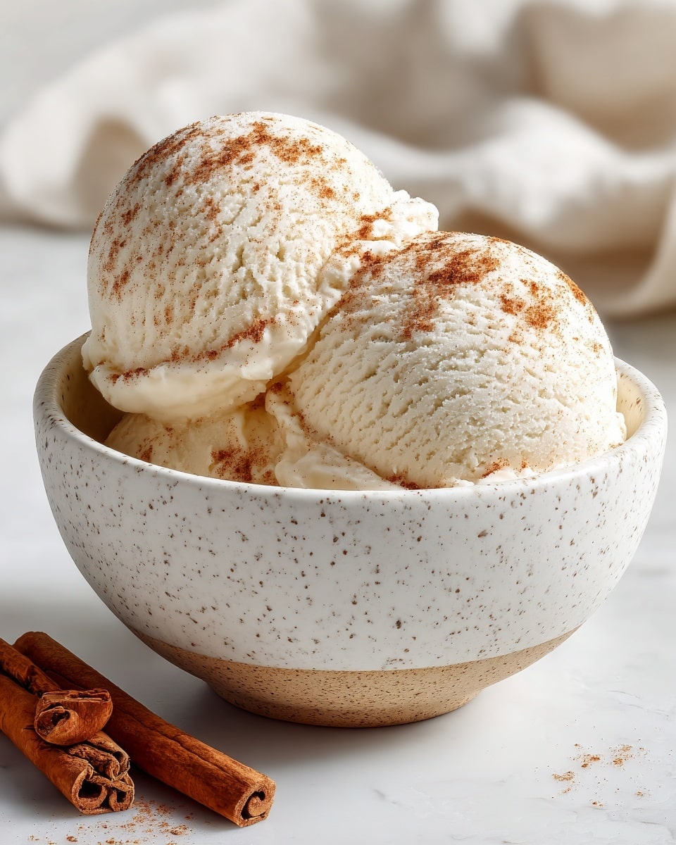 A white speckled bowl filled with a large scoop of creamy vanilla ice cream, textured with soft swirls and small air pockets, topped lightly with a sprinkle of cinnamon powder. The bowl sits on a white marbled surface, with a folded white cloth to the left and some brown cinnamon sticks blurred in the background, creating a cozy and inviting scene. photo taken with an iphone --ar 4:5 --v 7